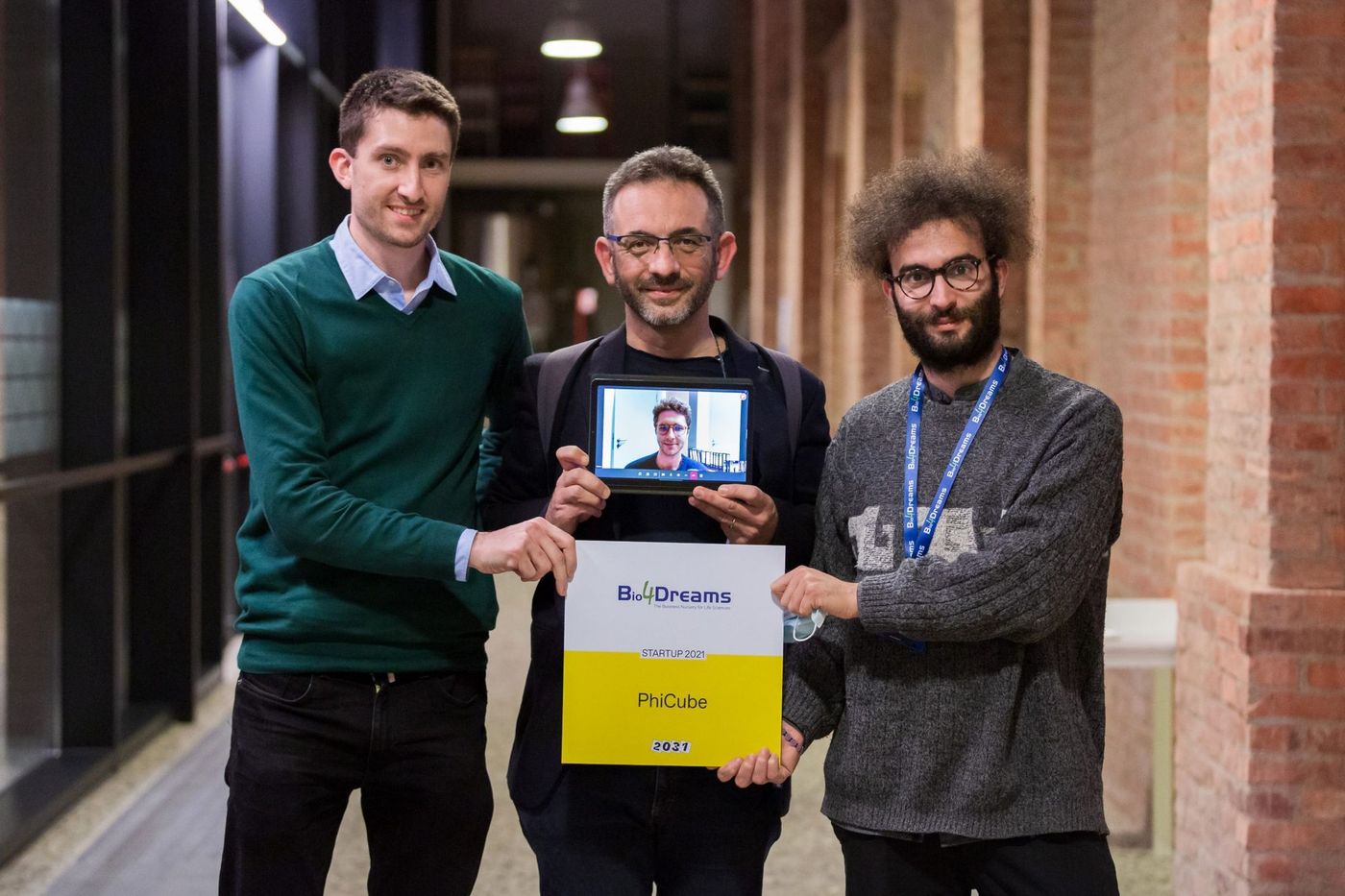 Drei Teammitglieder halten ein Bio4Dreams-Preisschild mit der Aufschrift „PhiCube“ und ein Tablet, auf dem ein per Videoanruf zugeschalteter Kollege zu sehen ist.