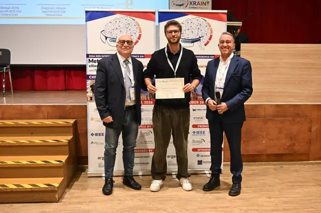 Matteo Lavit Nicora hält auf der Bühne der IEEE MetroXRAINE 2026 die Urkunde, flankiert von zwei Vertretern der Konferenz, vor den Bannern der Veranstaltung.