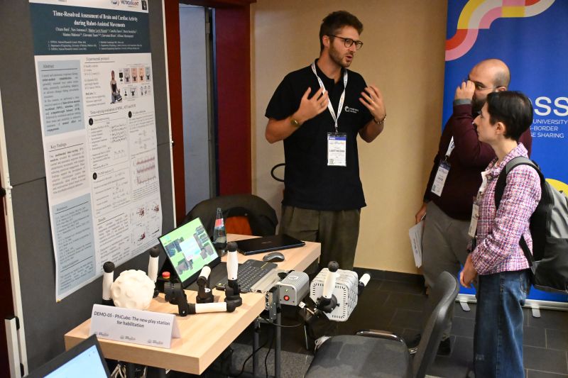 At the PhiCube booth, Matteo Lavit Nicora talks with two visitors beside a desk holding the robotic unit, its grips and a laptop running a game; the research poster hangs on the left.