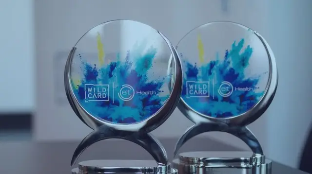 Two glass EIT Health Wild Card trophies with a blue and yellow splash design stand on a table.