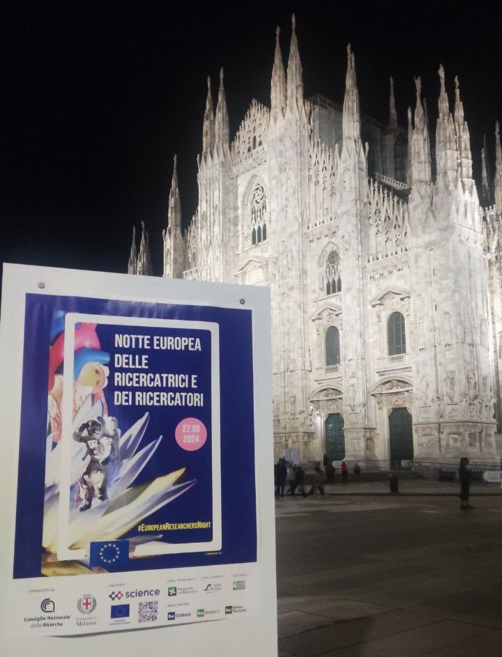A poster for the European Researchers’ Night stands in Piazza Duomo at night, with the illuminated Milan Cathedral behind it.