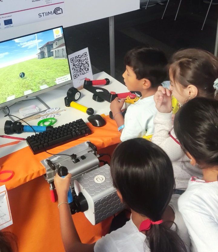 A group of children gather around the PhiCube device and its screen at an outdoor demonstration table.