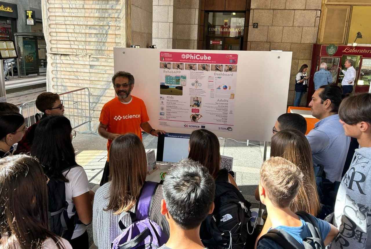 A presenter in an orange Science t-shirt explains a PhiCube poster to a group of students outdoors in Milan.
