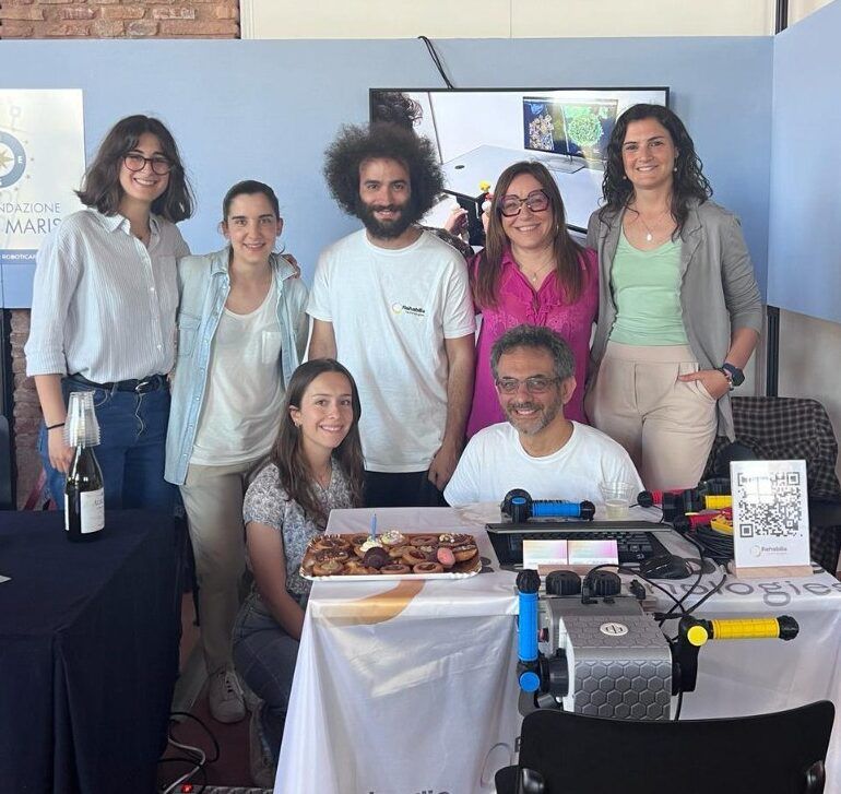 L’équipe de Rehabilia pose sur le stand de la Fondazione Stella Maris au Festival della Robotica, avec des pâtisseries et PhiCube sur la table.