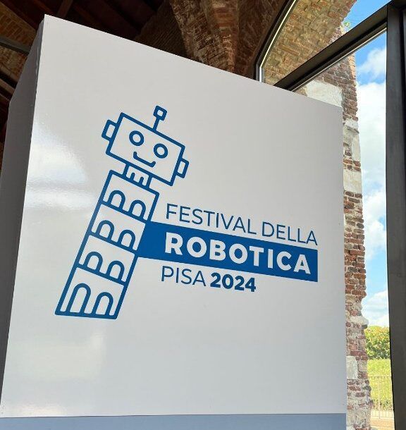 Un panneau portant le logo du Festival della Robotica Pisa 2024, un robot de dessin animé souriant, devant un mur de briques.