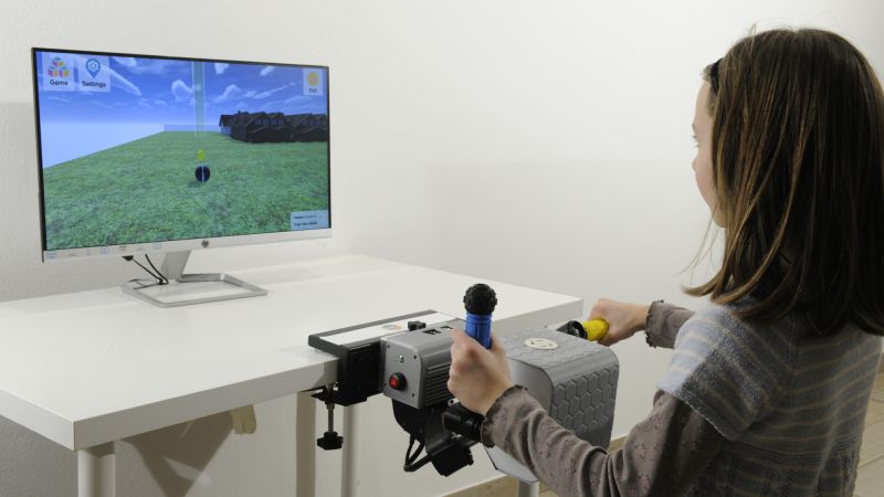 Une fillette assise à un bureau tient une poignée de PhiCube dans chaque main, devant un écran où tourne un jeu.