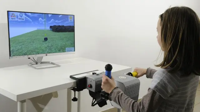 A child sits at a desk holding one PhiCube handle in each hand, with a game running on the monitor in front of her.