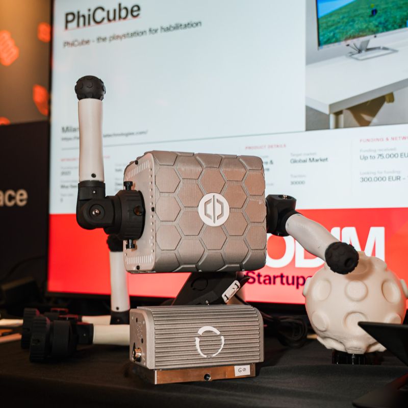 PhiCube expuesto en el estand de Rehabilia en PODIM 2026, con sus empuñaduras, su núcleo robótico y una pantalla que muestra el menú de juegos de la plataforma.