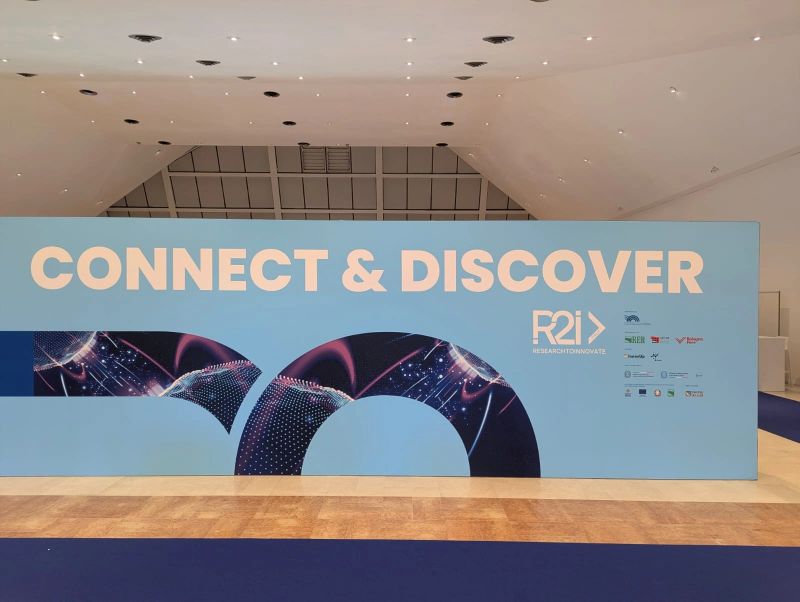 Eine Wand mit der Aufschrift „Connect & Discover“ und dem Logo von R2I – Research to Innovate im Ex GAM in Bologna.