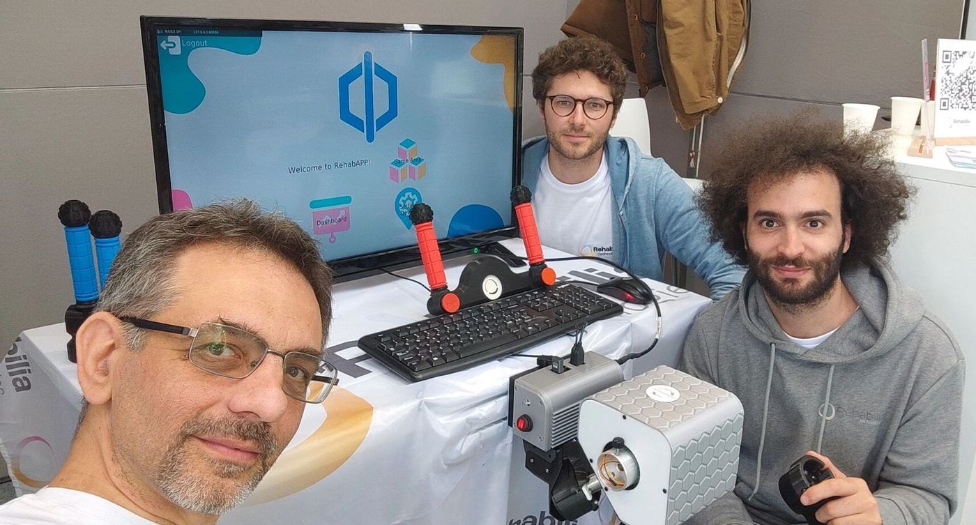 Die drei Gründer von Rehabilia machen am Stand auf der Exposanità ein Selfie; auf dem Tisch stehen PhiCube und ein Bildschirm mit der RehabAPP.