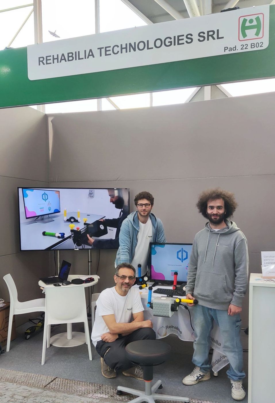 Rehabilia auf der Exposanità 2024 Drei Teammitglieder posieren am Stand von Rehabilia Technologies auf der Exposanità, unter dem Firmenschild und Bildschirmen, auf denen PhiCube gezeigt wird.