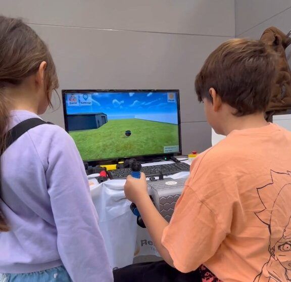 PhiCube auf der Exposanità 2024 Zwei Kinder spielen abwechselnd mit den Griffen von PhiCube vor einem Bildschirm mit einem 3D-Therapiespiel.