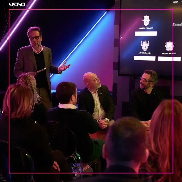 A speaker addresses a panel and audience at the WeRo Studio robotics event, with neon lighting and panellist names shown on screen behind him.